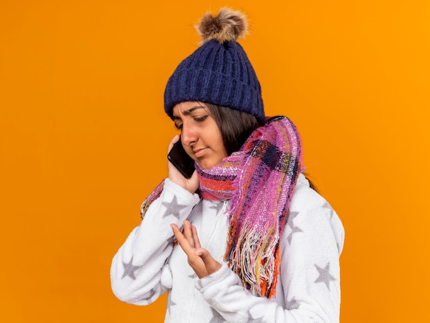 Niña enferma molesta con sombrero de invierno con bufanda habla por teléfono aislado sobre fondo amarillo