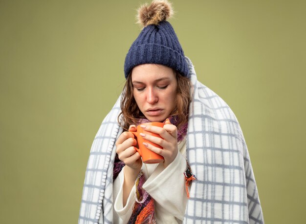 Niña enferma molesta vistiendo túnica blanca y gorro de invierno con bufanda envuelta en cuadros sosteniendo y mirando una taza de té aislado en verde oliva