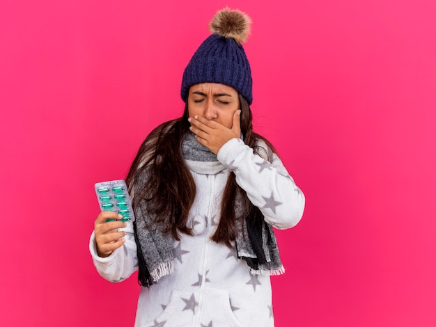 Niña enferma con los ojos cerrados con sombrero de invierno con bufanda sosteniendo pastillas cubiertas con la boca dolorida de la mano aislada en rosa