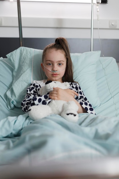 Niña enferma bajo tratamiento usando un tubo de oxígeno mientras sostiene un oso de peluche. Niño enfermo descansando solo en la cama del paciente del centro de salud infantil mientras tiene un oso de peluche.