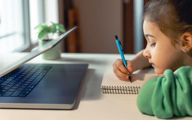 Una niña escribe en un cuaderno mientras se sienta frente a una computadora portátil