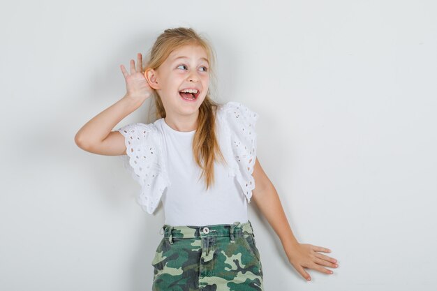 Niña escuchando algo con la mano sobre la oreja en camiseta blanca