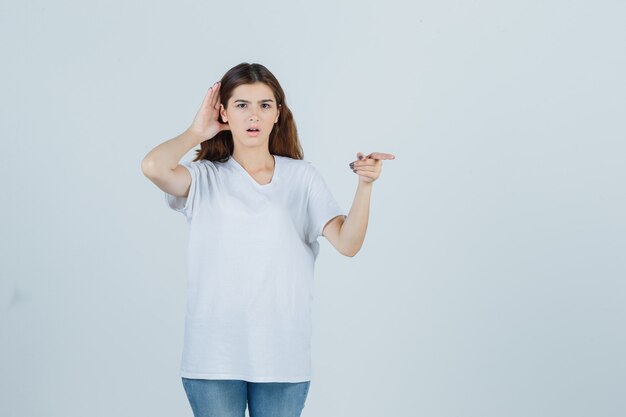 Niña escuchando una conversación privada, apuntando hacia otro lado con una camiseta blanca y mirando sorprendido. vista frontal.