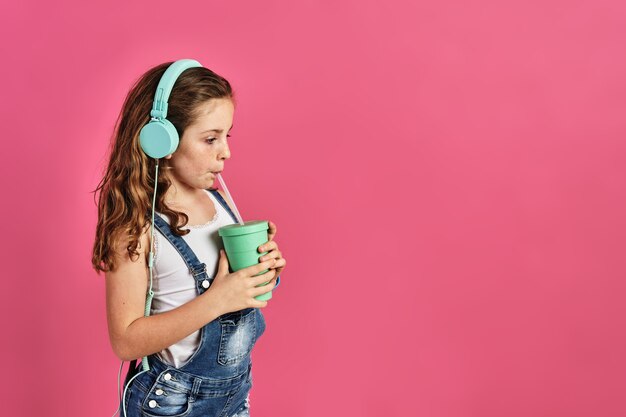 Niña escuchando música con auriculares y bebiendo un jugo en una pared rosa