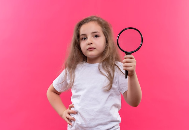 niña de la escuela con camiseta blanca sosteniendo lupa en la pared rosa aislada