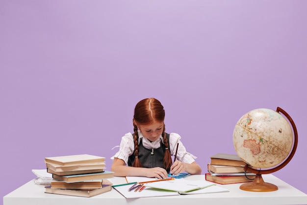 Niña de la escuela con dos trenzas rojas en blusa blanca y vestido de verano se sienta a la mesa con libros y globo sobre fondo lila