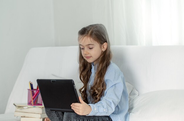 Foto gratuita una niña de escuela primaria se sienta en casa en el sofá con una computadora portátil en una lección en línea durante la cuarentena debido a la pandemia de coronavirus.