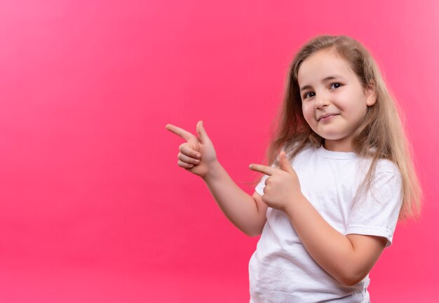 Niña de la escuela sonriente con camiseta blanca apunta al lado sobre fondo rosa aislado