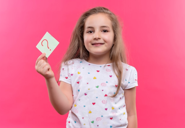 Niña de la escuela sonriente con camiseta blanca con signo de interrogación de papel sobre fondo rosa aislado