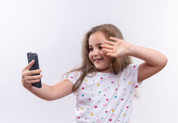 Niña de la escuela sonriente vistiendo camiseta blanca habla por teléfono sobre fondo blanco aislado