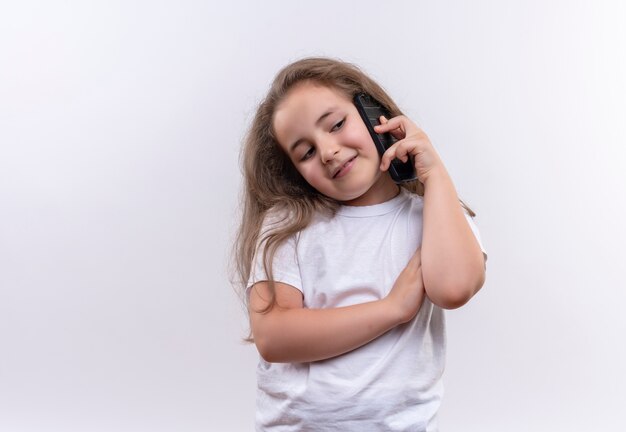 Niña de la escuela sonriente vistiendo camiseta blanca habla por teléfono sobre fondo blanco aislado