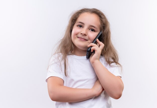 Niña de la escuela sonriente vistiendo camiseta blanca habla por teléfono sobre fondo blanco aislado