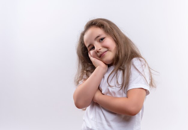 Niña de la escuela sonriente vistiendo camiseta blanca puso su mano en la mejilla sobre fondo blanco aislado