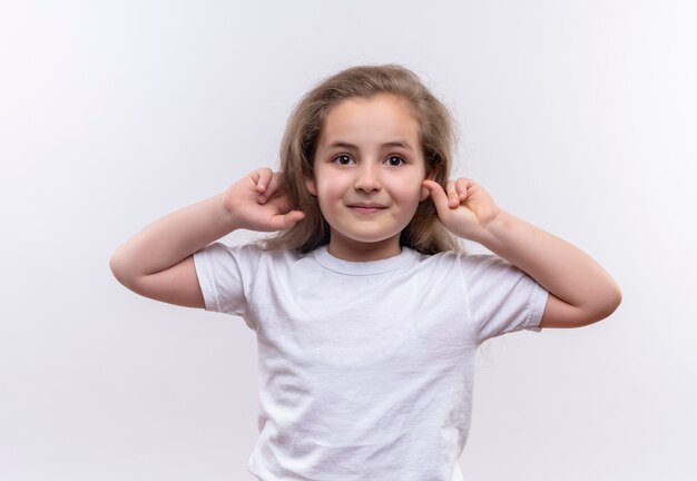 Niña de la escuela sonriente vistiendo camiseta blanca puso sus manos alrededor de las orejas sobre fondo blanco aislado