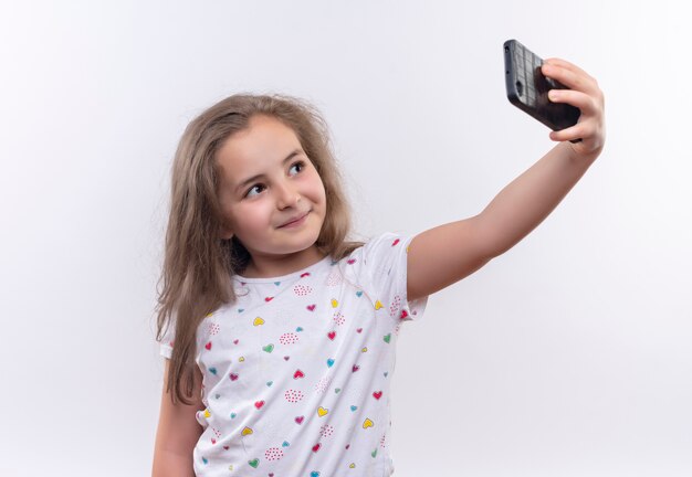 Niña de la escuela sonriente vistiendo camiseta blanca tomando selfie sobre fondo blanco aislado