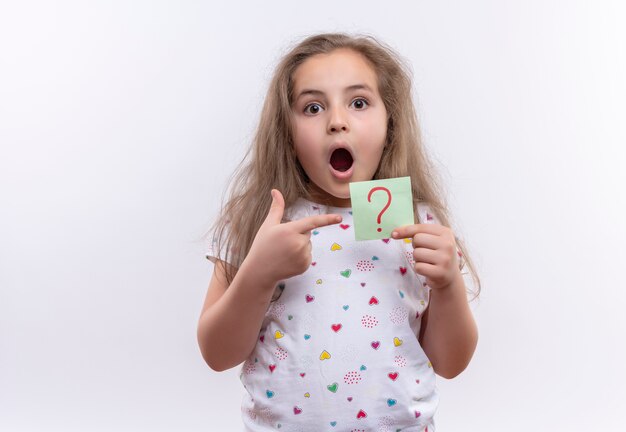 Niña de la escuela sorprendida con camiseta blanca apunta al signo de interrogación de papel sobre fondo blanco aislado