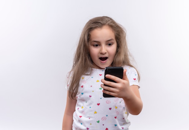 Niña de la escuela sorprendida con camiseta blanca mirando por teléfono sobre fondo blanco aislado