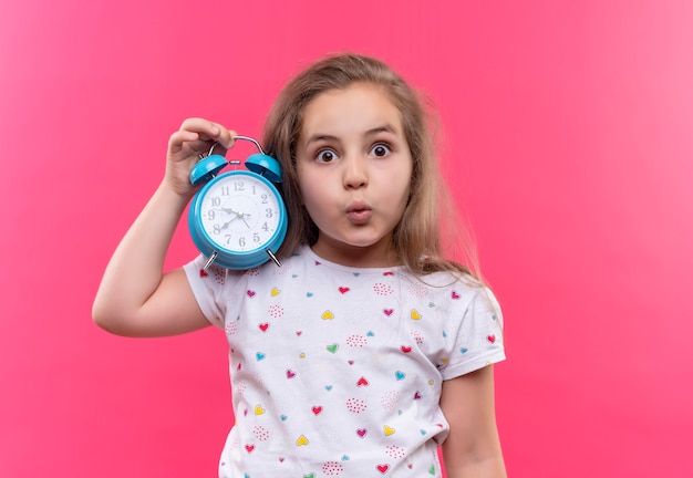 Foto gratuita niña de la escuela sorprendida con camiseta blanca con reloj despertador sobre fondo rosa aislado