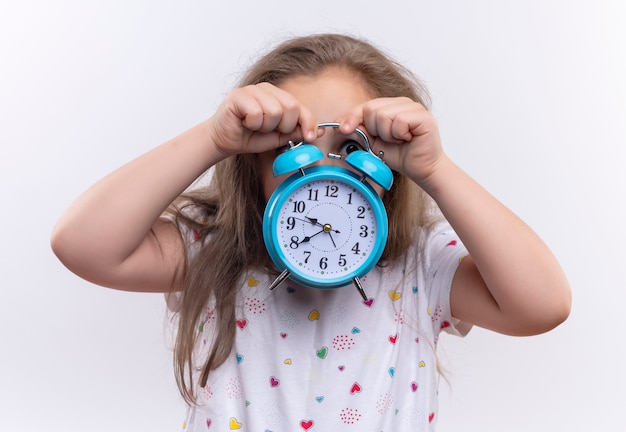 Niña de la escuela vistiendo camiseta blanca con la cara cubierta con reloj despertador en la pared blanca aislada