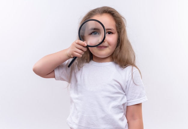 Niña de la escuela vistiendo camiseta blanca mirando a través de la lupa en la pared blanca aislada