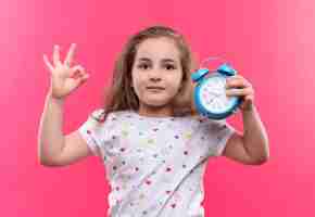 Foto gratuita niña de la escuela vistiendo camiseta blanca con reloj despertador mostrando gesto okey en pared rosa aislada