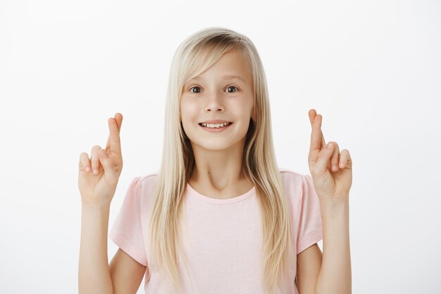 La niña espera que santa visite su casa con un gran regalo. Sonriendo soñadora hija joven con cabello rubio, levantando las manos con los dedos cruzados y sonriendo con asombro y deseo, esperando o pidiendo deseo