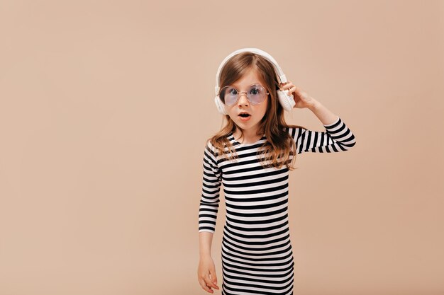 Niña con estilo sorprendida con vestido despojado y vidrio redondo de moda escuchando música y posando para la cámara