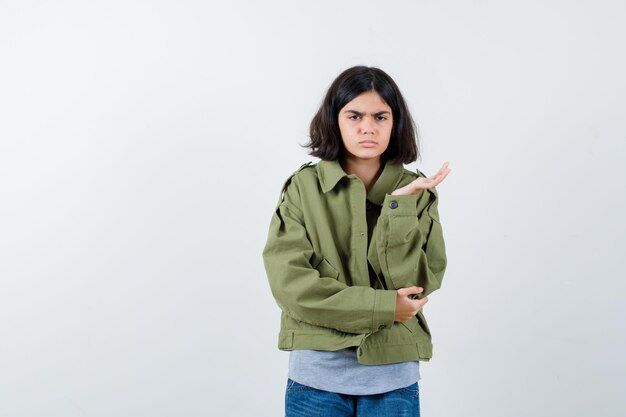 Niña estirando la mano en gesto de interrogación en abrigo, camiseta, jeans y mirando vacilante, vista frontal.