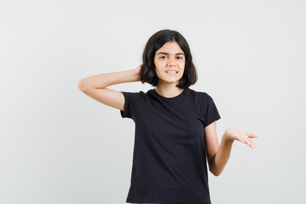 Niña estirando la mano de manera interrogante en camiseta negra y mirando alegre, vista frontal.