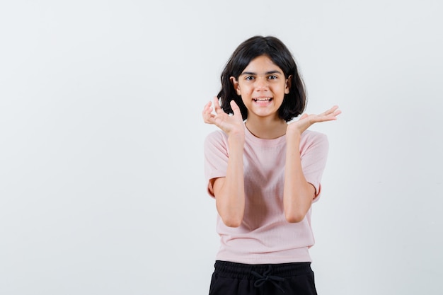 Niña estirando las manos como presentando algo en camiseta rosa y pantalón negro y mirando feliz