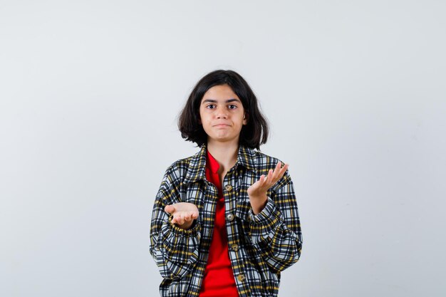 Niña estirando las manos en forma de interrogación en camisa a cuadros y camiseta roja y mirando perplejo, vista frontal.