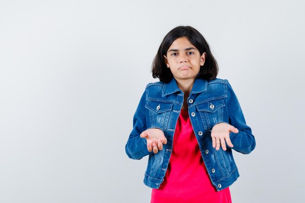 Niña estirando las manos en forma de interrogación en camiseta roja y chaqueta de jean y mirando desconcertado