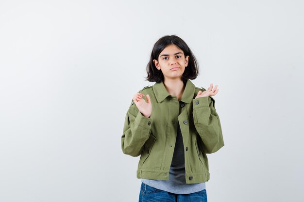 Niña estirando las manos de manera interrogante en suéter gris, chaqueta caqui, pantalón de mezclilla y mirando desconcertado, vista frontal.