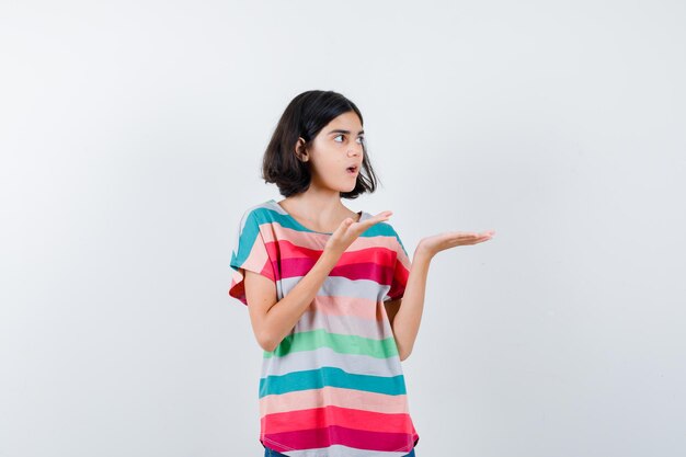 Niña estirando las manos sosteniendo algo en camiseta, jeans y mirando sorprendido, vista frontal.