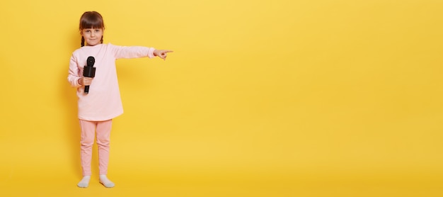 Foto gratuita niña europea con micrófono mira a la cámara mientras sostiene el micrófono, señala con el dedo índice a un lado en el espacio vacío para publicidad o promoción, vocalista encantador presenta algo en la pared amarilla