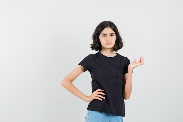 Niña extendiendo la palma a un lado en camiseta negra, pantalones cortos y mirando ansiosa. vista frontal.