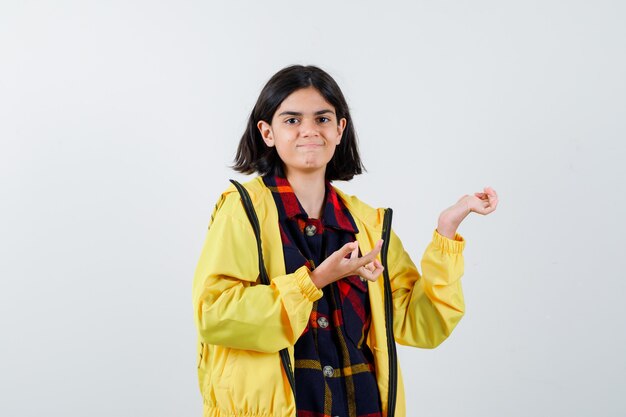 Niña extendiendo las palmas en camisa a cuadros, chaqueta y mirando confiado