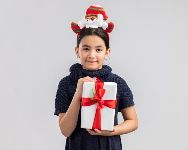 Niña feliz y alegre en vestido de punto con corbata roja con divertido borde navideño en la cabeza sosteniendo el regalo de Navidad mirando con una sonrisa en la cara