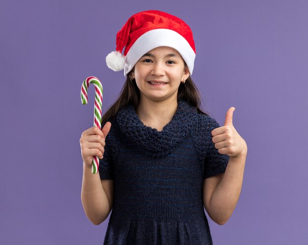 Niña feliz y alegre en vestido de punto con gorro de Papá Noel sosteniendo bastón de caramelo con una sonrisa en la cara mostrando los pulgares para arriba de pie sobre la pared púrpura