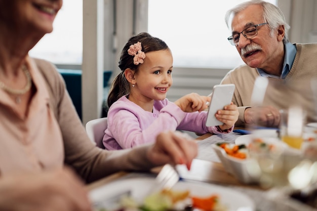 Niña feliz almorzando con sus abuelos y usando un teléfono inteligente en la mesa del comedor