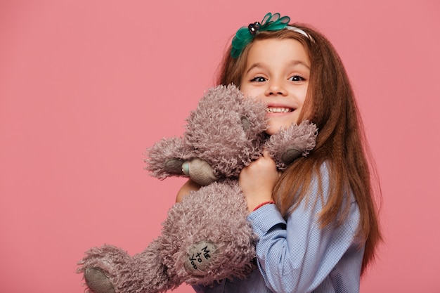 Niña feliz con cabello castaño largo sonriendo jugando con su adorable osito de peluche