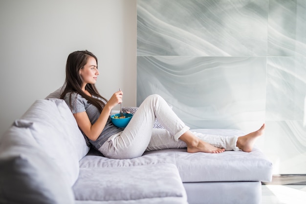 Niña feliz comiendo cereales con frutas de un tazón sentado en un sofá en casa