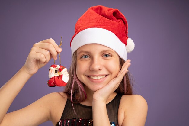 Niña feliz y complacida en vestido de fiesta brillante y gorro de Papá Noel con juguetes de Navidad mirando a la cámara sonriendo alegremente de pie sobre fondo púrpura
