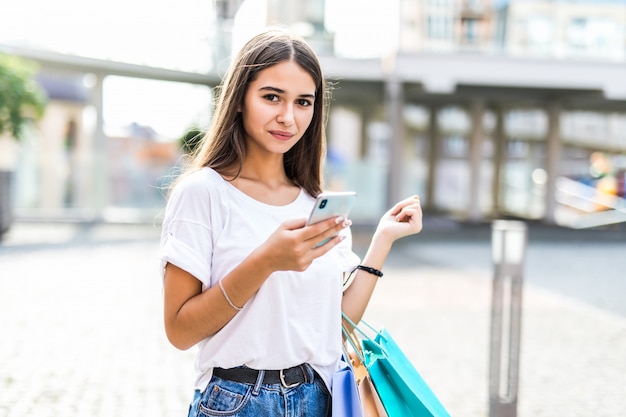 Niña feliz en compras saliendo del centro comercial con bolsas y mirando el teléfono.