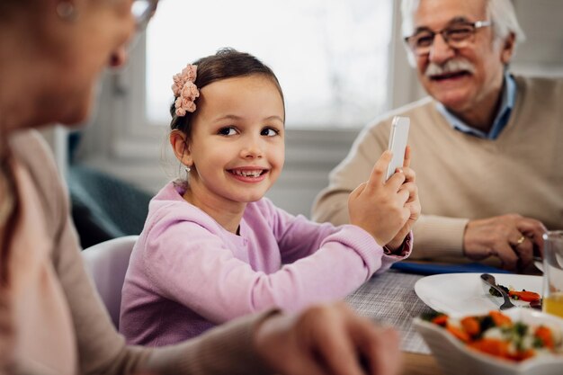 Niña feliz comunicándose con sus abuelos mientras usa el teléfono celular durante el almuerzo en la mesa del comedor