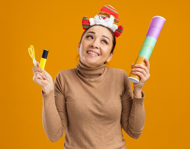 Niña feliz en divertido borde navideño en la cabeza sosteniendo petardos tijeras y pegamento