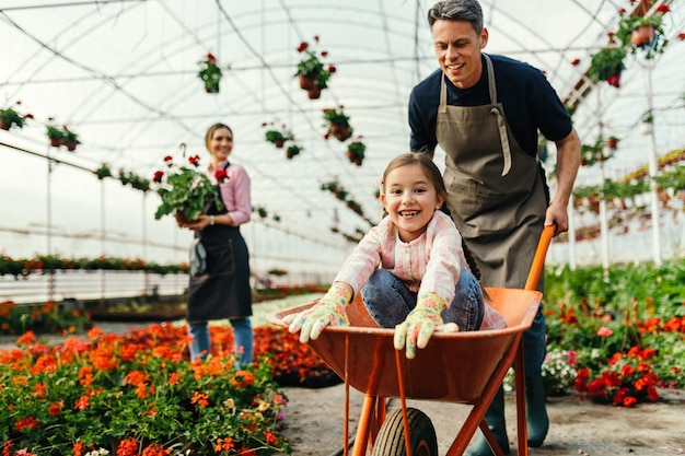 Una niña feliz divirtiéndose mientras su padre la empuja en una carretilla en el vivero de plantas La madre está trabajando en segundo plano