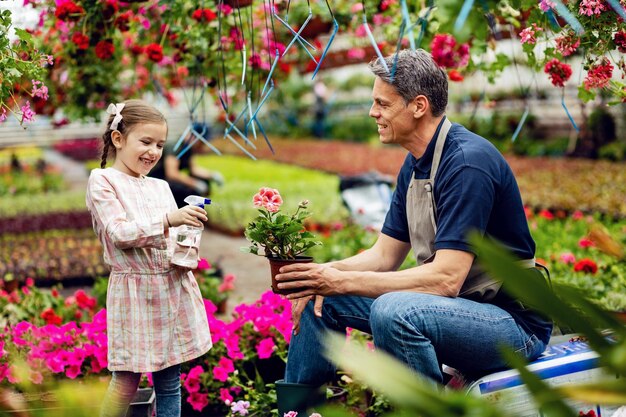 Niña feliz divirtiéndose con su padre mientras usa una botella de spray y riega flores en macetas en el vivero de plantas