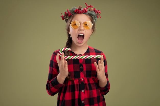 Niña feliz y emocionada con corona de navidad en vestido comprobado sosteniendo bastón de caramelo mirando a la cámara gritando de pie sobre fondo verde