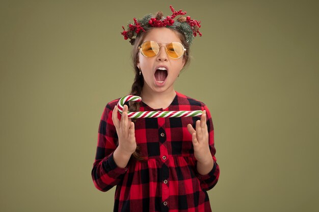 Niña feliz y emocionada con corona de navidad en vestido comprobado sosteniendo bastón de caramelo mirando a la cámara gritando de pie sobre fondo verde